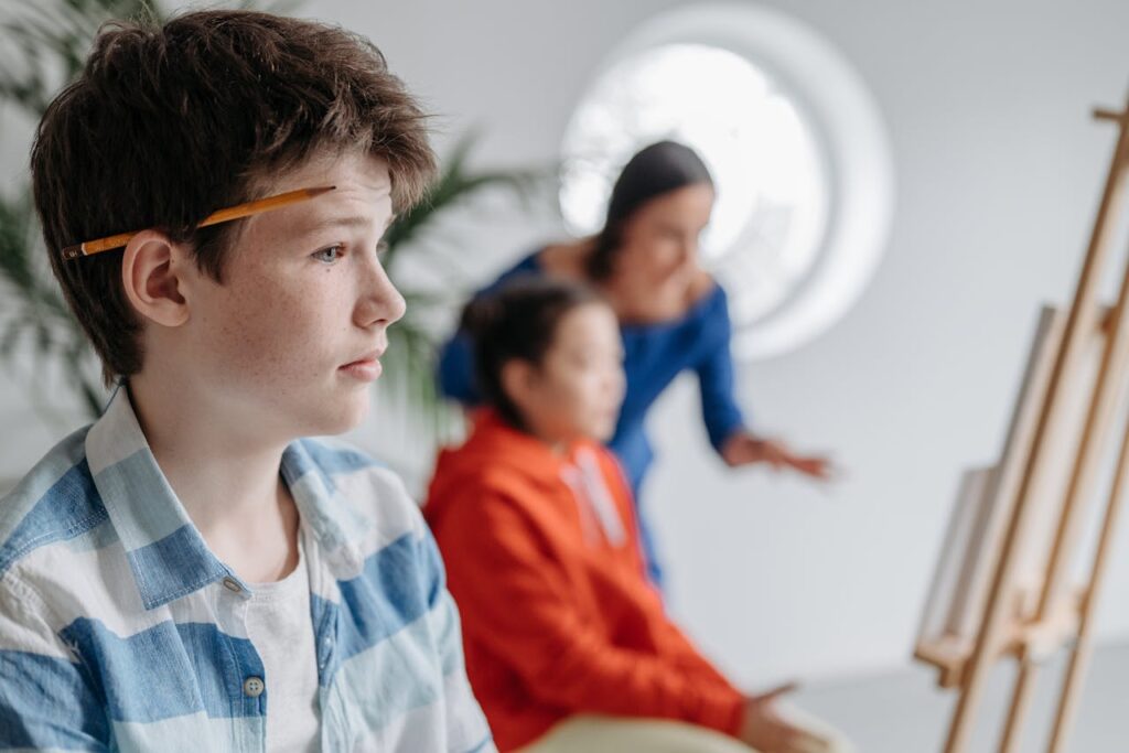 Boy with a Pencil behind the Ear and Teacher Talking to a Girl in the Background during an Art Class